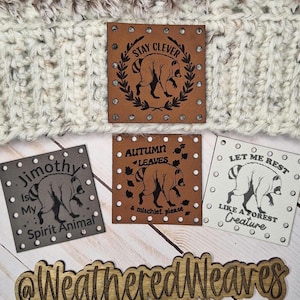 Op de afbeelding: Een crème en bruine gehaakte muts met textuur, voorzien van een vierkante lichtbruine leren patch met een wasbeer en de tekst Stay Clever. Drie extra vierkante patches met wasbeerillustraties en tekst, plus een houten bordje met de tekst WeatheredWeaves.