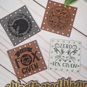 May include: Four square leather tags with fox designs and text. The tags are in shades of grey, brown, and white. One tag says "Stay Clever", another says "Cozy Days", and two say "Zero Fox Given" and "Iuro Fox Given".