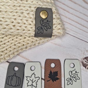 Pode incluir: Chapéu de malha creme com uma etiqueta de couro cinza com um desenho de folha e um fecho de pressão dourado. Abaixo, quatro etiquetas de couro com desenhos temáticos de outono, incluindo abóboras e folhas, em várias cores.