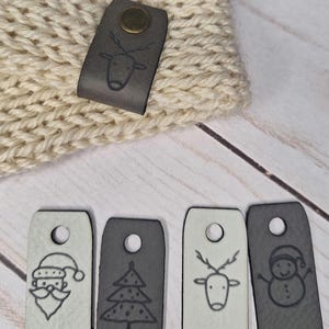 Pode incluir: Chapéu de malha creme com uma etiqueta de couro cinza com um design de rena. Várias etiquetas de couro cinza e branco com ilustrações temáticas de Natal, incluindo o Papai Noel, uma árvore de Natal, renas e um boneco de neve.
