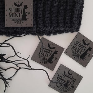 Pode incluir: Quatro patches quadrados de tecido cinza com costuras pretas e texto preto. Os patches apresentam uma silhueta de gato preto e um desenho de teia de aranha. O texto nos patches diz "Spooky Mini", "Spooky Dad", "Spooky Mama" e "Spooky Vibes".