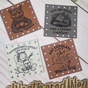 May include: Four square leather patches with illustrations. One features a raccoon with a mug and the text "Warm Wishes." Another depicts a fox with the text "Let Me Rest Like a Forest Creature." The third has a fox in a chair with the text "Coverstimulated and Over It." The last shows a fox with a mug and the text "Hot Chocolate is Like a Warm Hug."