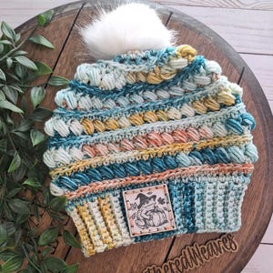 May include: A hand-crocheted beanie in shades of teal, yellow, and peach, topped with a fluffy white pom-pom. The hat features a textured stitch pattern and a small leather tag with a cartoon frog and pumpkin. The text "Weathered Weaves" is visible.