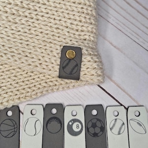May include: Cream-colored knit beanie with a baseball emblem on a leather tag. Includes a selection of sports-themed leather tags featuring basketball, tennis, golf, 8-ball, soccer, baseball, and football designs. A cozy accessory for sports enthusiasts.