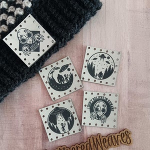 May include: Four metal square patches with black and white alien and UFO designs. The patches have a dotted border and are on a wooden surface. The text on one patch reads "I don't believe in humans". The text on another patch reads "Area 51". A wooden sign with the text "@WeatheredWeaves" is in the bottom right corner of the image.