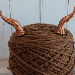 May include: A ball of brown yarn with two copper-colored horns attached. The horns are curved and textured. The image also includes the text "NEW COLORS!"