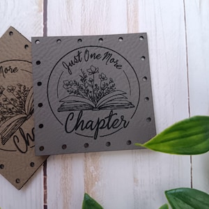 May include: Gray leather patch with a circle design featuring a book with flowers and the text "Just One More Chapter".