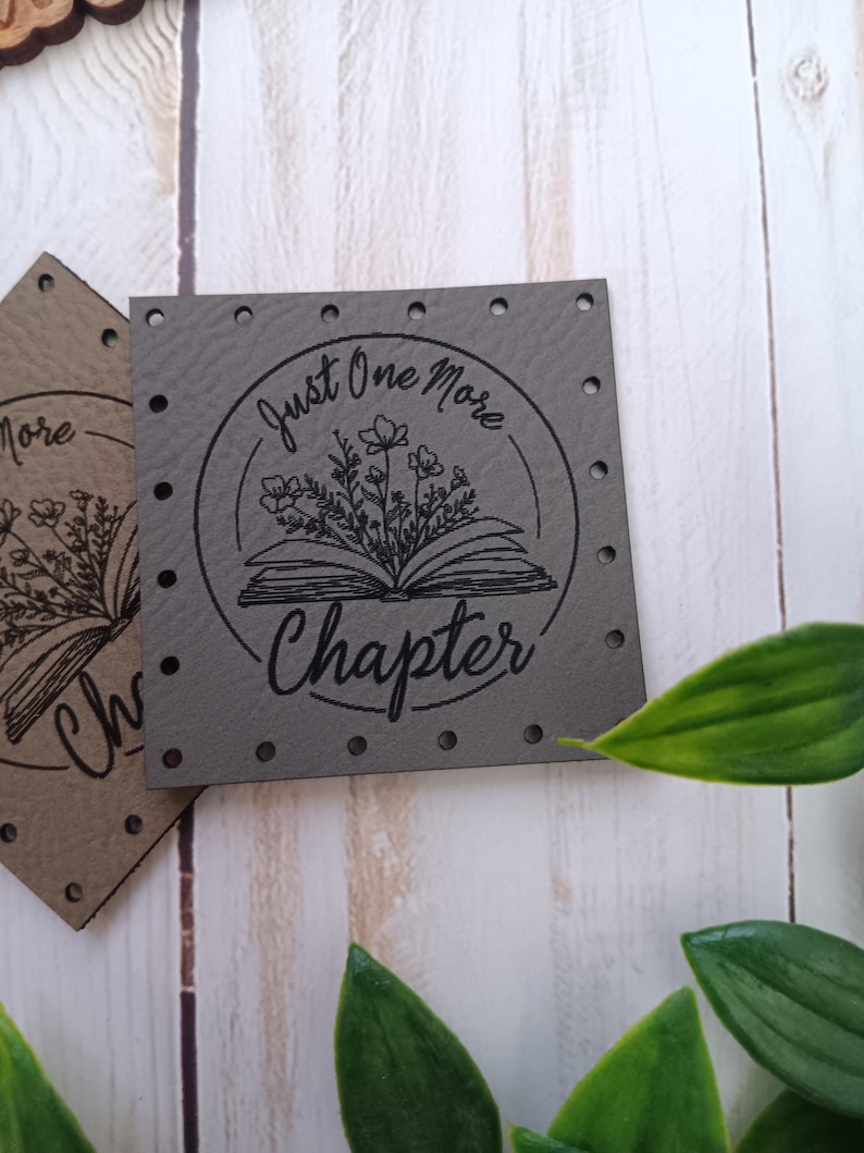 May include: Gray leather patch with a circle design featuring a book with flowers and the text "Just One More Chapter".