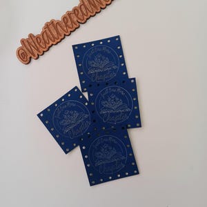 May include: Four blue square fabric bookmarks with a white design of an open book with flowers and the text "Just One More Chapter".