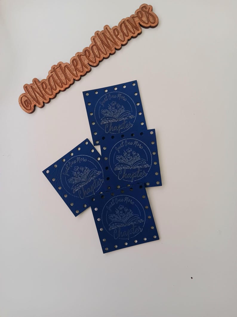 May include: Four blue square fabric bookmarks with a white design of an open book with flowers and the text "Just One More Chapter".