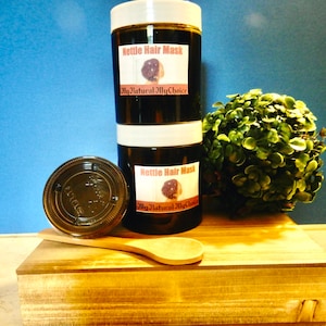 May include: Two glass jars of dark brown nettle hair mask stacked on top of each other. The jars have white lids and labels that say "Nettle Hair Mask" and "My Natural My Choice". The jars are sitting on a wooden box with a wooden spoon.