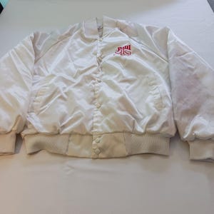 May include: A white satin bomber jacket with a ribbed collar, cuffs, and hem. The jacket features a button-up front and two front pockets. The text "Paul & Lisa" is embroidered in red on the upper left chest.