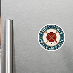 May include: A circular sticker featuring a red ball of yarn with two crossed knitting needles in the center. The design is enclosed in a teal border with the text "KNITTING CIRCLE" at the top and "ANTI-FASCIST SOCIETY" at the bottom in white.