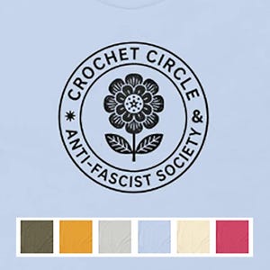 May include: A light blue t-shirt featuring a black circular graphic on the chest. The design includes a central flower illustration surrounded by the text "CROCHET CIRCLE & ANTI-FASCIST SOCIETY". Below the graphic is a row of six color swatches.