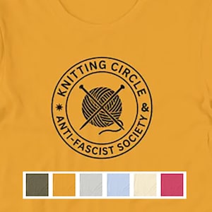 May include: A mustard yellow short-sleeved t-shirt featuring a black circular graphic on the chest. The design includes a ball of yarn with crossed knitting needles, surrounded by the text KNITTING CIRCLE & ANTI-FASCIST SOCIETY. A color palette bar is below.