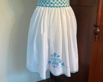 May include: A white cotton apron with a blue and green gingham ribbon belt and blue floral embroidery. The apron has a scalloped lace trim.