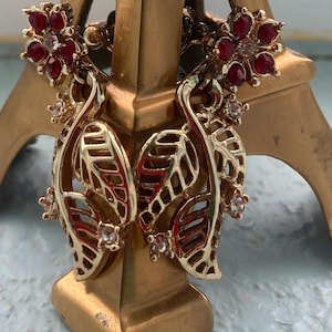 May include: Gold-toned earrings with a leaf design and red and clear gemstones. The earrings feature a floral cluster at the top and small gemstones throughout the leaf design. The earrings are displayed on a gold-colored stand.