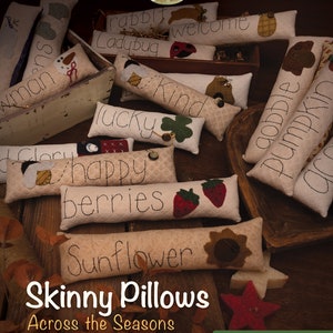 May include: A collection of handmade skinny pillows with various seasonal themes, including "rabbit", "welcome", "ladybug", "kind", "lucky", "happy", "berries", "sunflower", "gobble", "pumpkin", and "winter". The pillows are arranged in a wooden crate and on a wooden surface with autumn leaves. The text "Skinny Pillows Across the Seasons" is displayed at the bottom of the image, along with the text "Under the Woolen Willow".