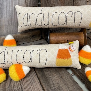 May include: Two decorative pillows with the words "candy corn" and "du corn" embroidered on them. The pillows are made of a beige fabric with a diamond pattern. There are several felt candy corn decorations on the wooden surface.