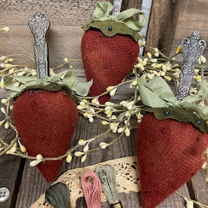Könnte beinhalten: Drei handgefertigte Erdbeer-Nadelkissen mit silbernen Löffelstielen, grünen Stoffblättern und grünem Band. Die roten Nadelkissen sind mit kleinen, dunklen Perlen verziert. Stickgarn, Spitze und Knöpfe sind ebenfalls sichtbar.