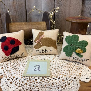 May include: Three small, square, cream-colored pillows with felt appliqués of a ladybug, rabbit, and clover. The ladybug is red and black, the rabbit is brown, and the clover is green. Each pillow has a word stitched on it.