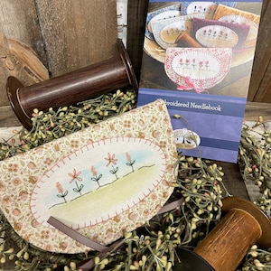 May include: A half-circle needle book with a floral pattern and embroidered flowers. The needle book is made of fabric and has a brown ribbon tied around it. The needle book is sitting on a wooden surface with a spool of thread and a garland of green and pink berries.