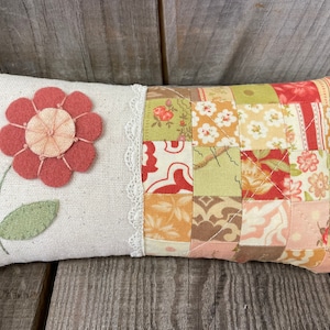 May include: A rectangular decorative pillow with a floral design. The left side features a large, felt flower in shades of pink and peach, with green leaves. The right side has a patchwork pattern of various floral fabrics. A white lace trim separates the two sides.