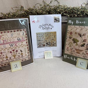 May include: Three cross-stitch pattern books are displayed, featuring intricate designs and text. The book covers showcase floral and alphabet patterns, with the text "Harriette Elizabeth Cox", "Barbara Ana Designs", and "Blackbird Designs". Small, decorative letter stamps are placed in front.