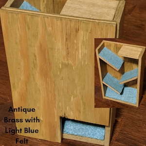 May include: Wooden dice tower with a light blue felt interior. The tower has a slanted design with multiple compartments for dice to roll down. The tower is made of natural wood with a light brown finish. The text "Antique Brass with Light Blue Felt" is visible on the front of the tower.