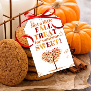 Op de afbeelding: Een wit cadeau-etiket met een rood lint eromheen, met de tekst "Just a little FALL TREAT for someone SWEET!" en een herfstboomillustratie. Het etiket ligt op een stapel peperkoekjes, twee mini-pompoenen, kaneelstokjes en steranijs op bruin perkamentpapier.