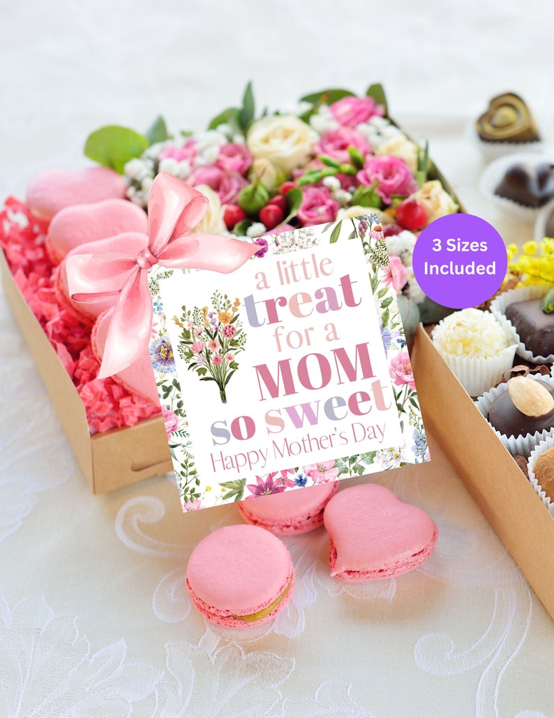 May include: A gift box filled with pink macarons and a floral arrangement. A card with the text "a little treat for a MOM so sweet Happy Mother's Day" is attached to the box with a pink ribbon. The box is sitting on a white tablecloth.