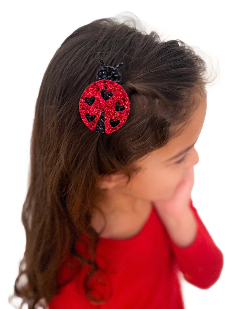 以下が含まれることがあります： 黒のディテールとグリッターが付いた赤いテントウムシのヘアクリップ。テントウムシは黒い輪郭と羽に黒いハートを持っています。