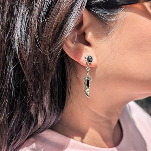 May include: Silver dangle earrings with black onyx stones. The earrings feature a feather design with a small round stone at the top.