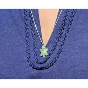 May include: A silver necklace with a small, carved, green turtle pendant. The turtle pendant is suspended from a delicate silver chain. The necklace is displayed against a blue background, with a scalloped trim.