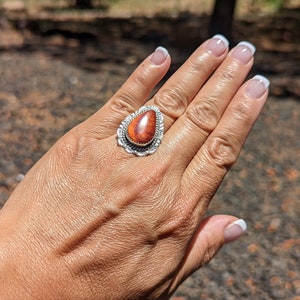 Puede incluir: Un anillo de plata con una piedra naranja en forma de lágrima engastada en un engaste de plata. La piedra tiene un patrón de mármol y está rodeada por un diseño de filigrana de plata.