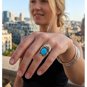 May include: A silver ring featuring a central oval turquoise stone surrounded by decorative metal scrollwork and leaf-like detailing. The ring is worn on a finger, accompanied by three plain silver bangles on the same wrist.