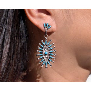 May include: A silver-toned earring with turquoise accents. The earring features an oval design with turquoise rods radiating outwards. A smaller cluster of turquoise stones is at the top. The earring is worn on a person's ear.