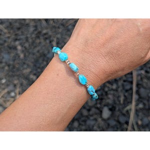 Puede incluir: Una pulsera de cuentas turquesa con detalles plateados. La pulsera presenta piedras de turquesa azul claro de forma ovalada, ensartadas con cuentas de metal plateado. La pulsera se lleva en la muñeca.