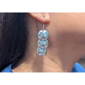 May include: Silver dangle earrings with three circular, textured elements. Each disc features a detailed sunburst pattern. The earrings are suspended from a simple hook, showcasing a classic and elegant design.
