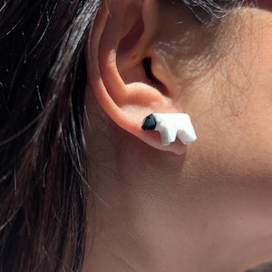 May include: White polar bear stud earrings with black details. The earrings are small, sculpted in a minimalist style, and are worn on the ear. The earrings are a unique accessory.