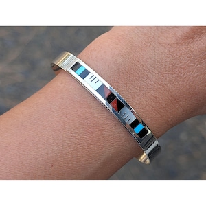 Puede incluir: Una pulsera de plata con diseños geométricos incrustados en negro, blanco, rojo y turquesa. La pulsera se muestra en una muñeca, mostrando su metal pulido y su diseño colorido. Un accesorio elegante.