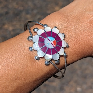 May include: A silver bracelet with a circular design featuring a red, white, and blue inlay. The center of the design is a red circle with white and blue accents. The bracelet is made of silver and has a floral design.