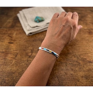 Könnte beinhalten: Ein Silberarmreif mit einer Kanaleinlage aus kleinen rechteckigen Segmenten aus Türkis, roter Koralle, schwarzem Onyx und gelber Muschel. Das Armband hat eine polierte Metalloberfläche und eine schmale, offene Bandkonstruktion.