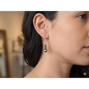 May include: Silver dangle earrings with three textured, round beads. The earrings are suspended from a simple hook and are shown on a person's ear. The beads have a slightly hammered appearance, reflecting light.