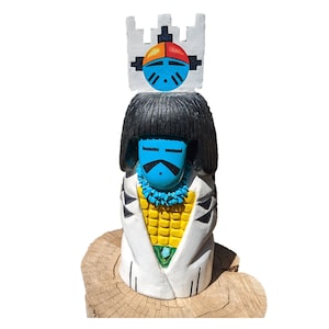 May include: Hand-carved wooden figurine of a Native American person wearing a white robe with a yellow and blue design. The figurine has a blue face with black eyes and a black mustache. The figurine is wearing a turquoise necklace and is standing on a wooden base.