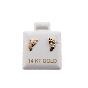 May include: A pair of gold-colored foot-shaped stud earrings on a white display card. The card has the text "14 KT GOLD" printed in gold. The earrings are small and detailed, with lines representing toes.