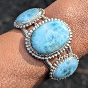 May include: A silver cuff bracelet featuring three oval-shaped, light blue larimar stones. Each stone is set within a beaded, silver frame. The bracelet is a piece of jewelry.
