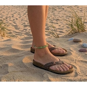 May include: A person wearing brown leather sandals with a buckle and a green beaded ankle bracelet. The sandals have a cork footbed. The person is standing on a sandy beach with some grass and small rocks.
