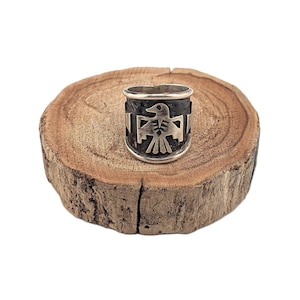 Op de afbeelding: Een zilveren ring met een gedetailleerd Native American thunderbird-ontwerp. De ring heeft een brede band met een donkere achtergrond en de thunderbird is prominent in het midden weergegeven. De ring ligt op een rustiek houten oppervlak.