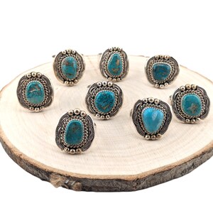May include: Eight turquoise rings with silver settings on a wooden surface. The rings feature oval-shaped turquoise stones with a dark blue and green color.